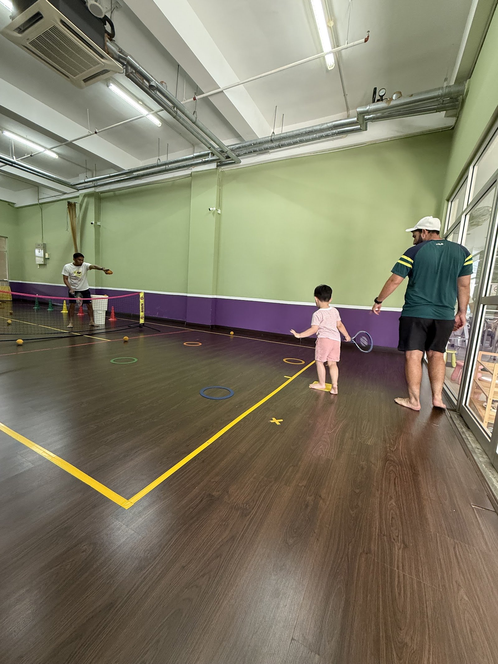 A Monster Tennis coach guiding a toddler with a racquet on the indoor court at Publika